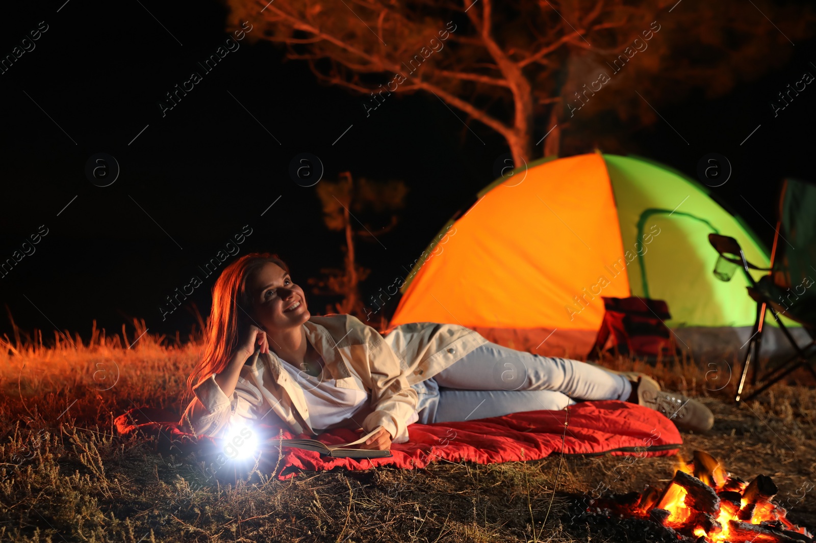 Photo of Young woman with flashlight and book near bonfire at night. Camping season