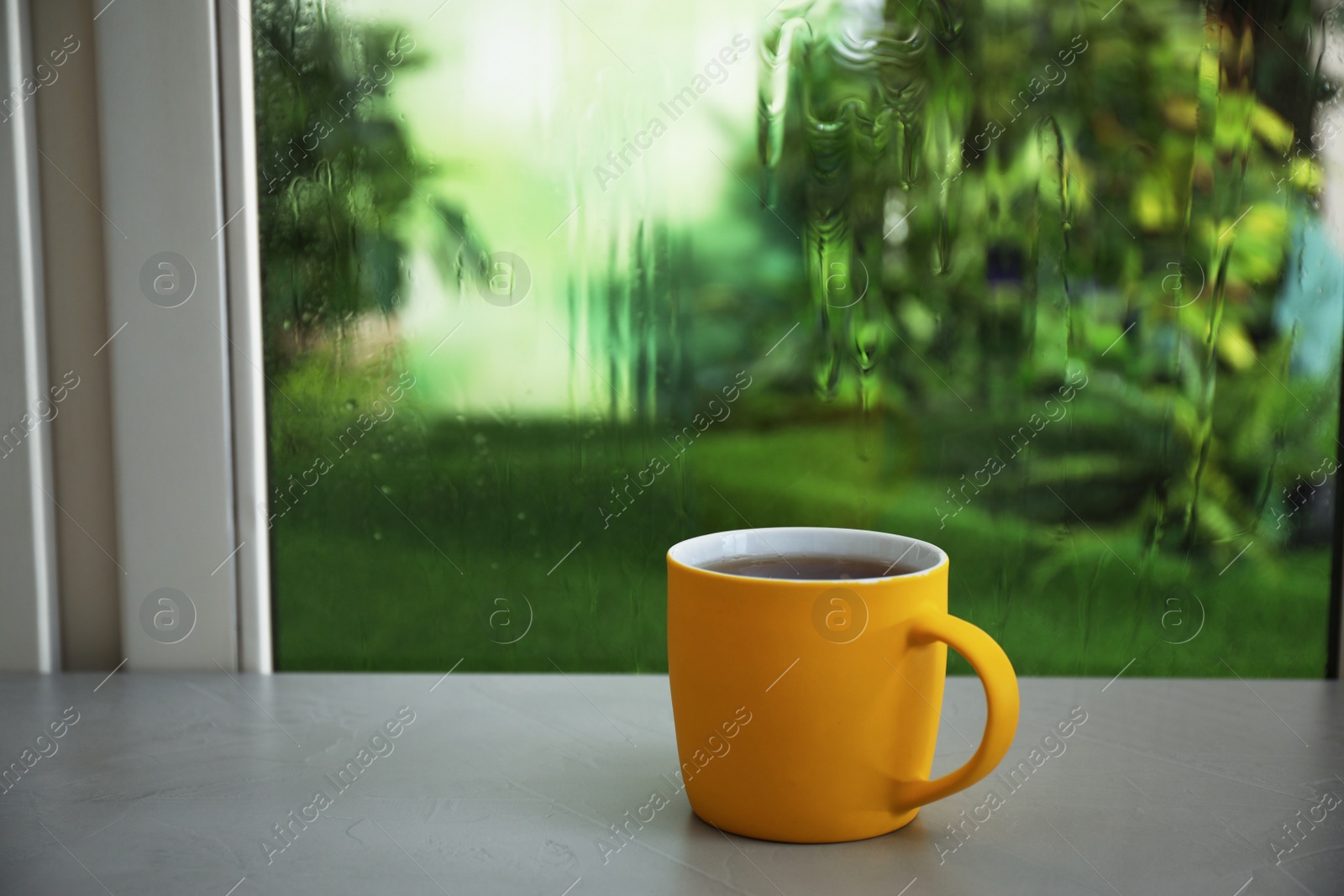 Photo of Cup of hot drink on windowsill against glass with rain drops, space for text