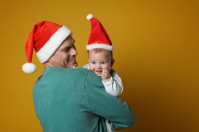 Photo of Happy father with cute baby wearing Santa hats on yellow background. Christmas season