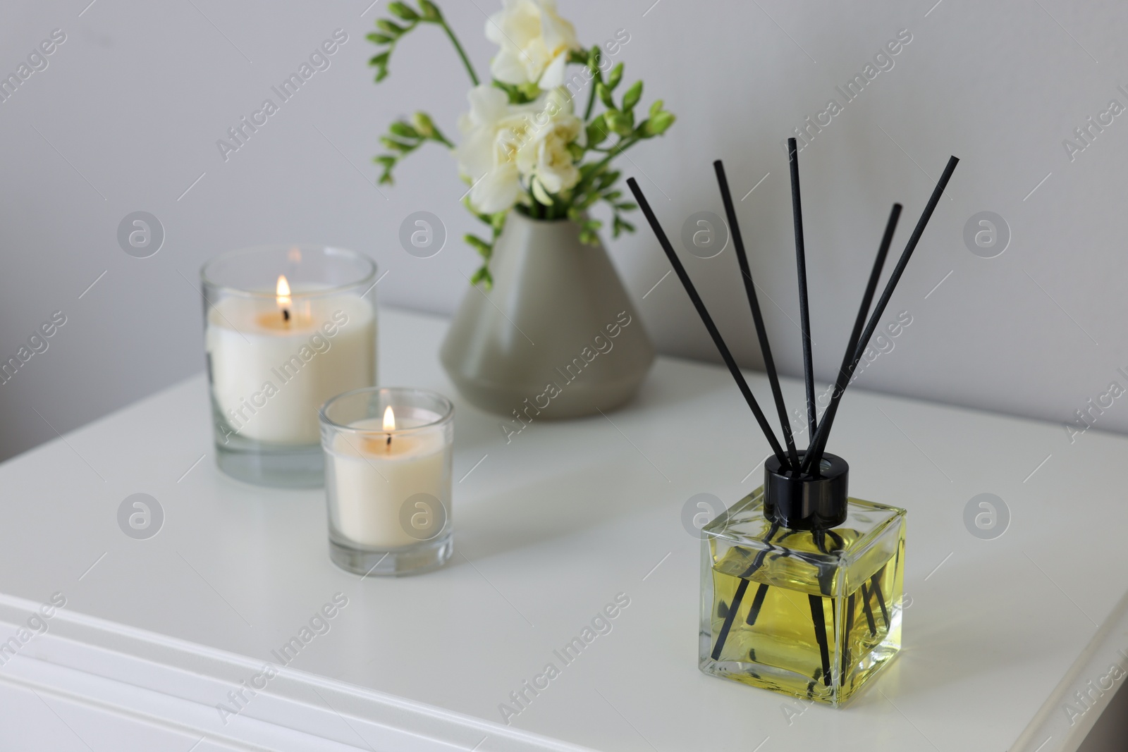 Photo of Aromatic reed air freshener, freesia flowers and candles on white bedside table indoors