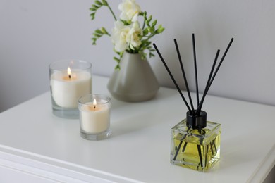 Photo of Aromatic reed air freshener, freesia flowers and candles on white bedside table indoors