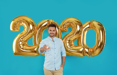 Happy young man with glass of champagne near golden 2020 balloons on turquoise background. New Year celebration