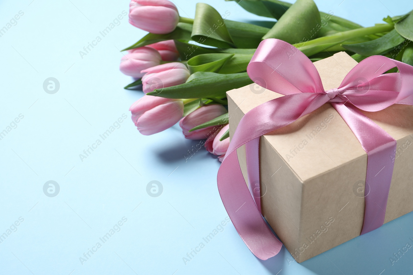 Photo of Beautiful gift box with bow and pink tulip flowers on light blue background, closeup. Space for text