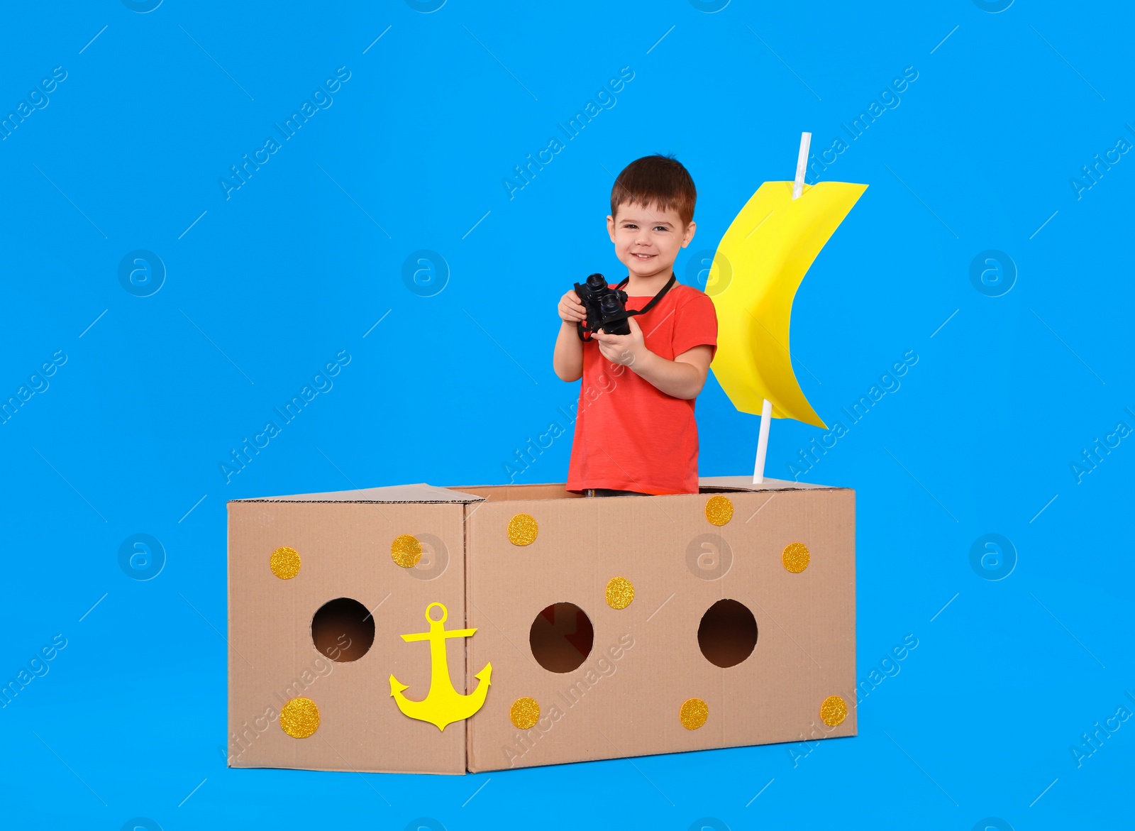 Photo of Cute little child playing with binoculars and cardboard ship on light blue background