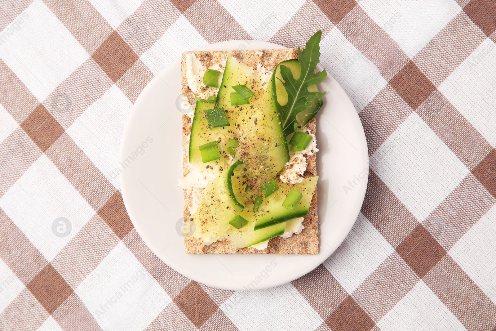 Photo of Fresh crunchy crispbread with cream cheese, cucumber, green onion and arugula on checkered tablecloth, top view