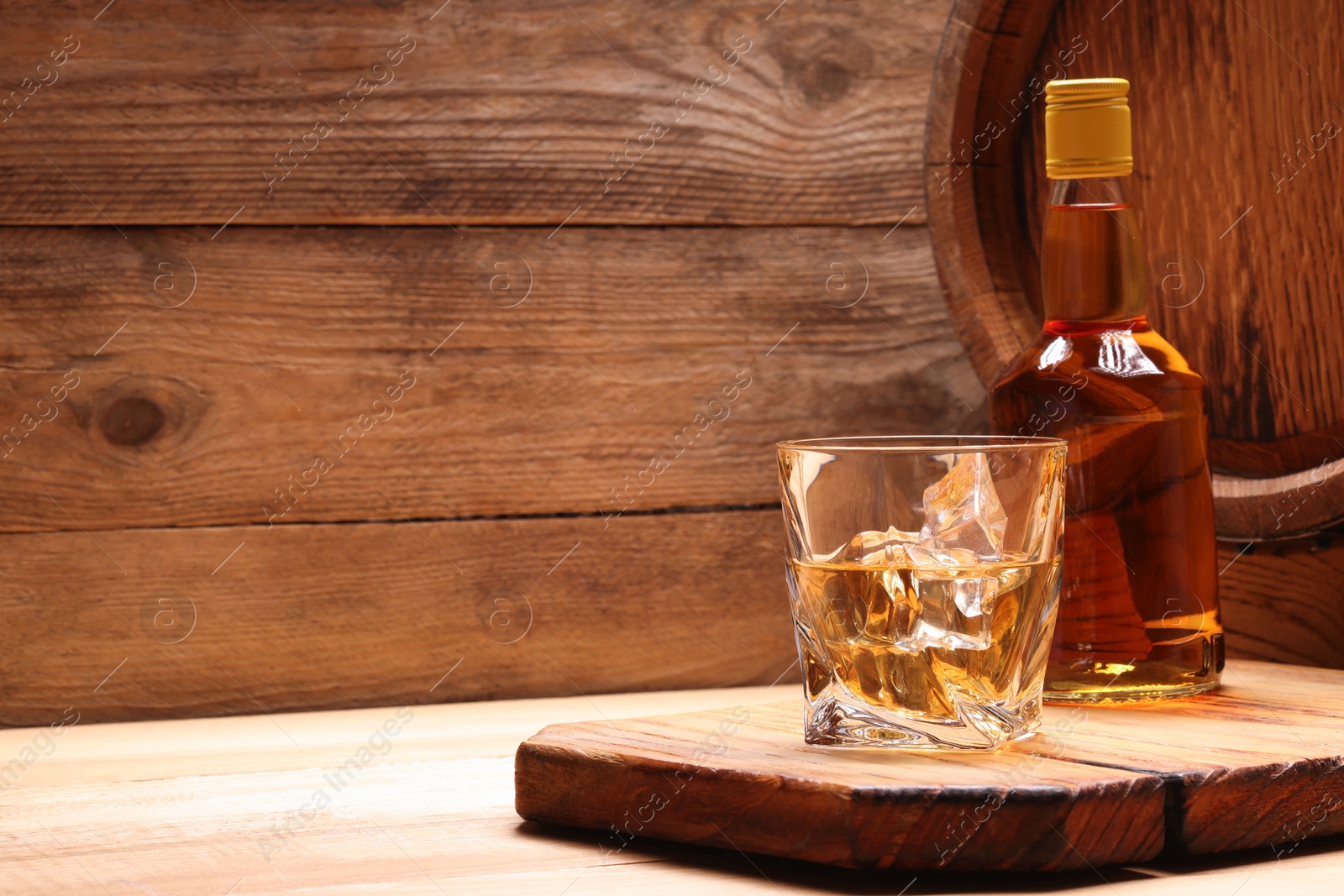 Photo of Whiskey with ice cubes in glass, bottle and barrel on wooden table, space for text