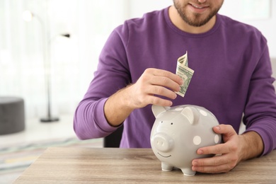 Young man putting money into piggy bank at table indoors. Space for text