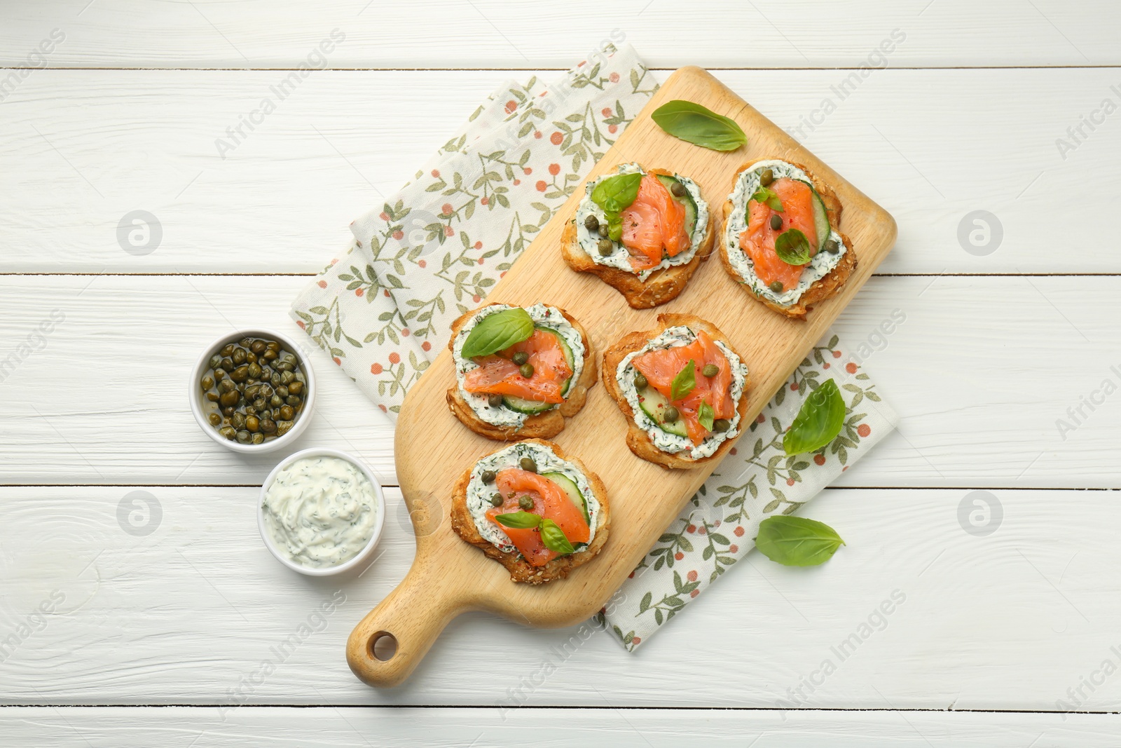 Photo of Tasty canapes with salmon, capers, cucumber and sauce on white wooden table, top view