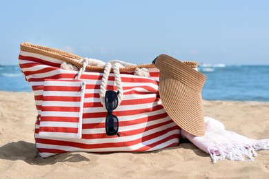 Photo of Stylish striped bag with visor cap, sunglasses and blanket on sandy beach near sea