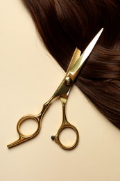 Photo of Professional scissors with brown hair strand on beige background, top view