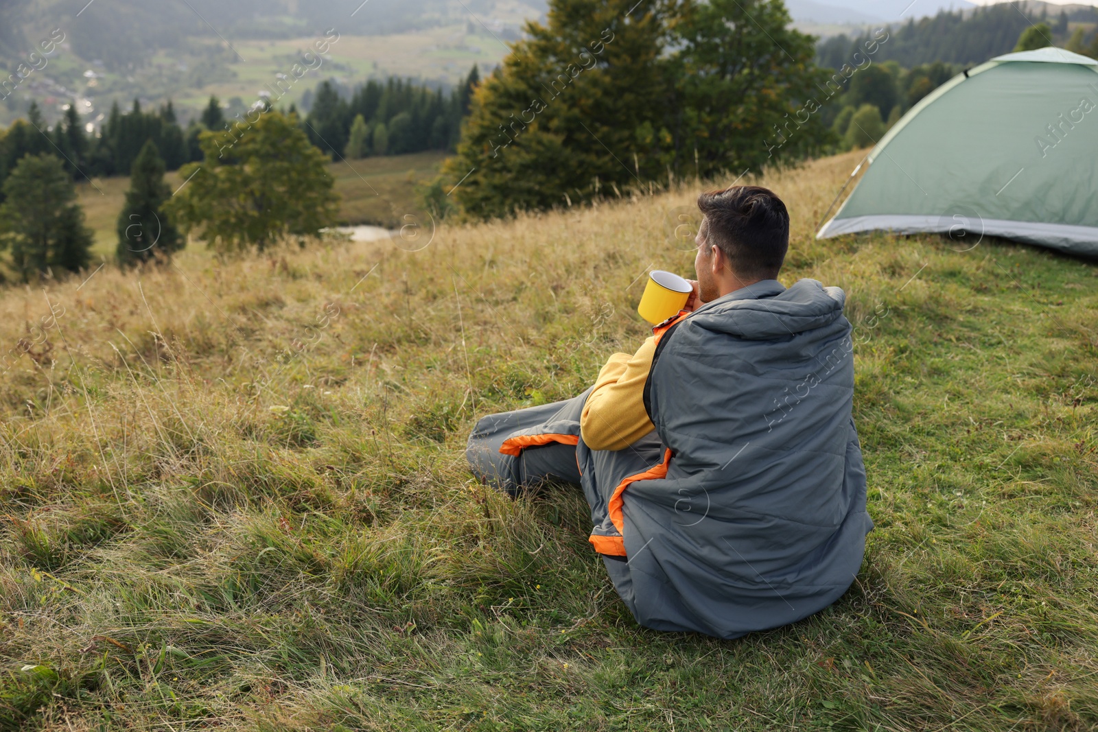 Photo of Man with cup of drink in sleeping bag enjoying mountain landscape, back view. Space for text