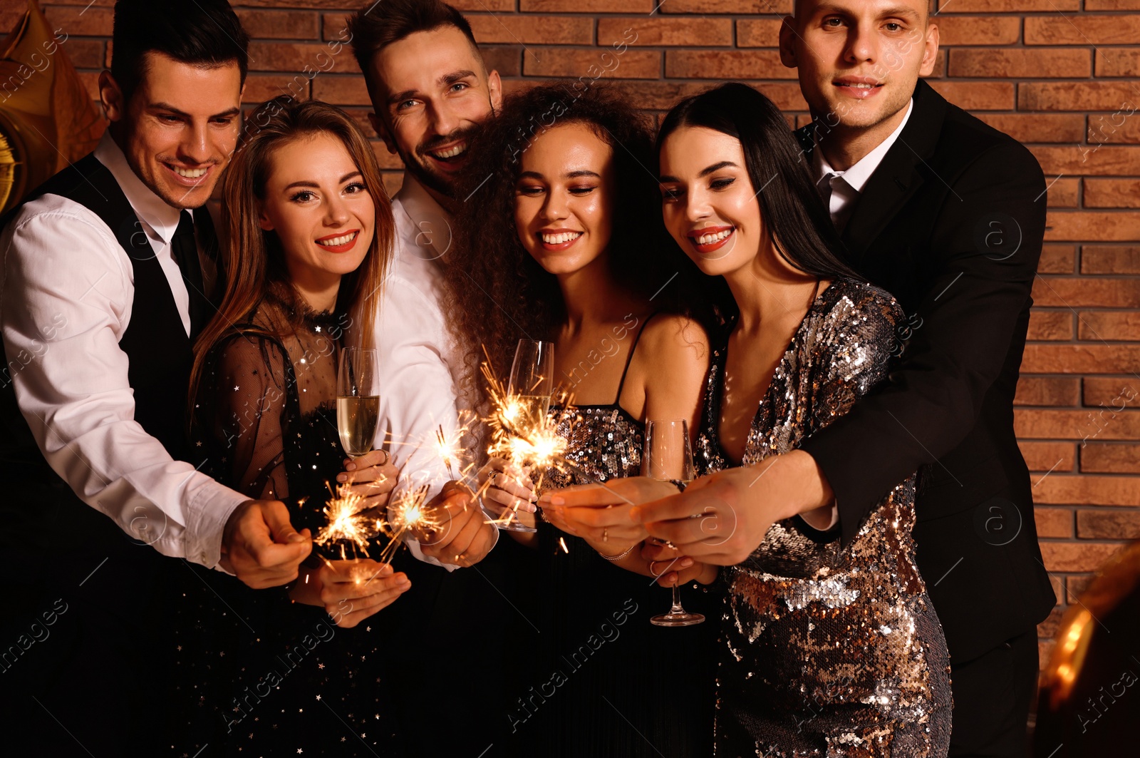 Photo of Happy friends with glasses of sparkling wine and sparklers celebrating New Year indoors