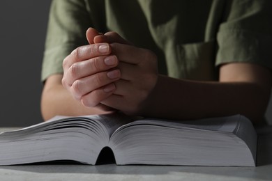 Woman holding hands clasped while praying over Bible at grey textured table, closeup