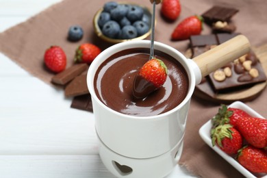 Photo of Dipping fresh strawberry in fondue pot with melted chocolate at white wooden table