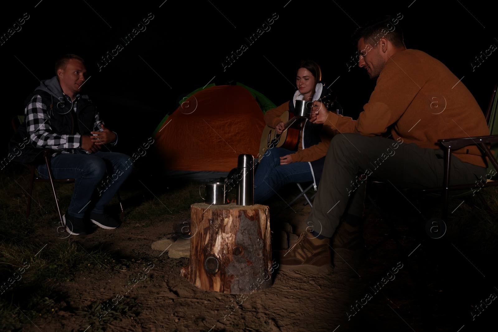 Photo of Group of friends with guitar near bonfire and camping tent outdoors at night