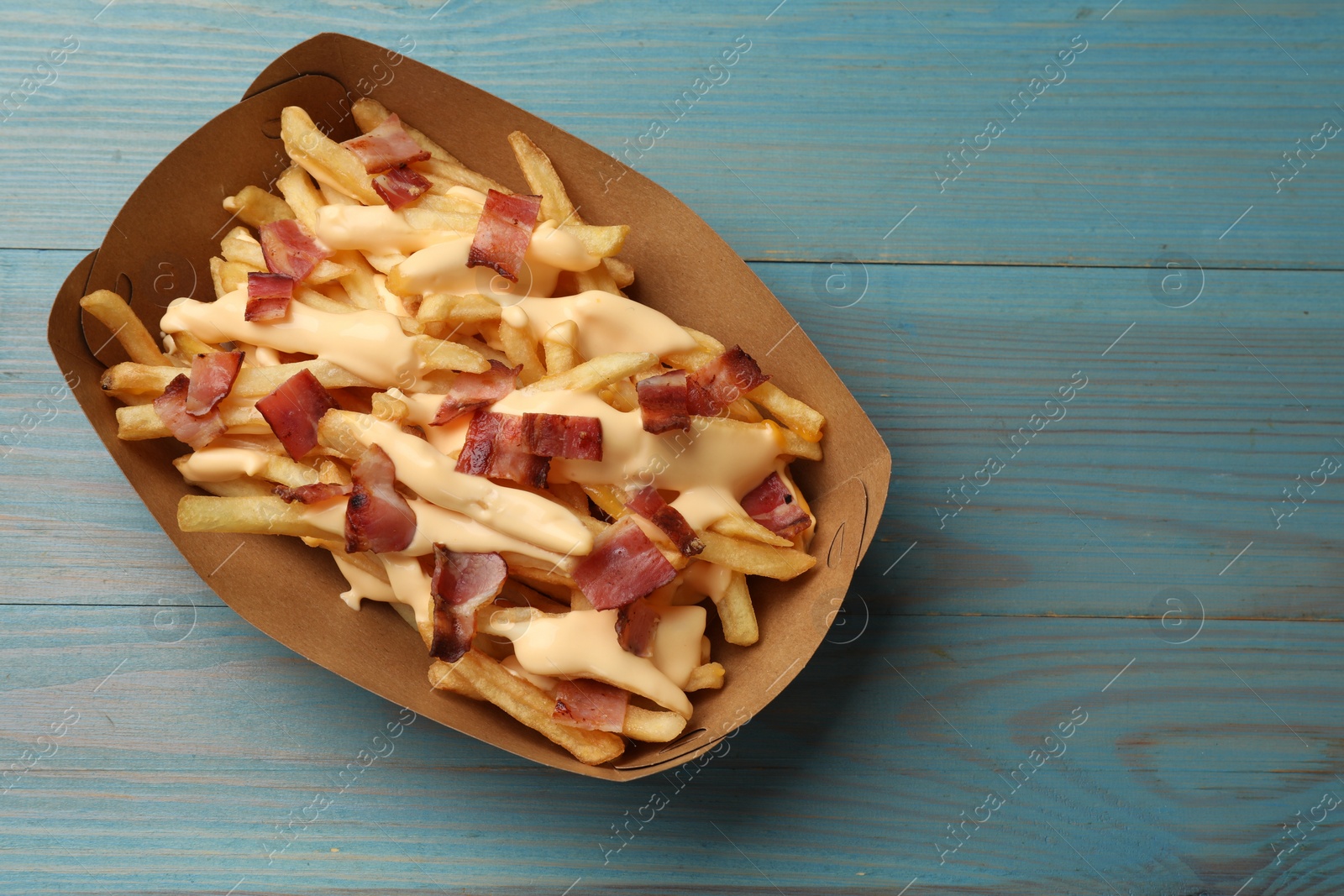 Photo of Tasty potato fries, cheese sauce and bacon in paper container on light blue wooden table, top view. Space for text