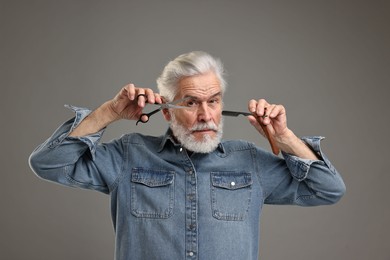 Senior man with mustache holding blade and scissors on grey background