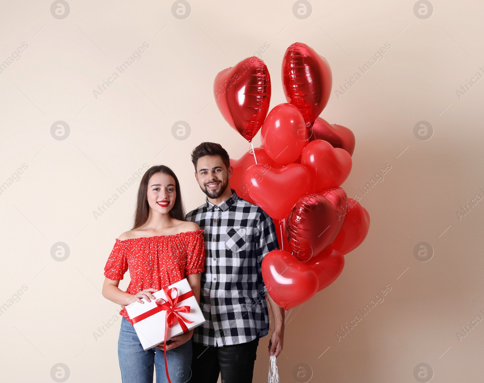 Photo of Happy young couple with gift box and heart shaped balloons on beige background. Valentine's day celebration