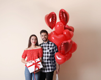 Photo of Happy young couple with gift box and heart shaped balloons on beige background. Valentine's day celebration