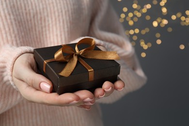 Photo of Christmas present. Woman holding gift box against grey background with blurred lights, closeup. Space for text