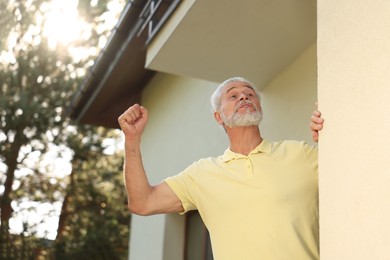 Photo of Angry senior man showing fist near house, low angle view. Annoying neighbour