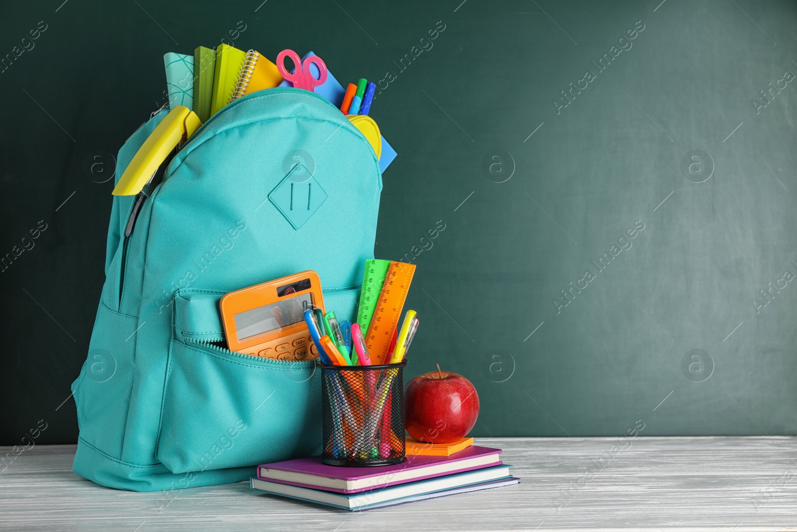 Photo of Stylish backpack with different school stationary on white wooden table at green chalkboard. Space for text