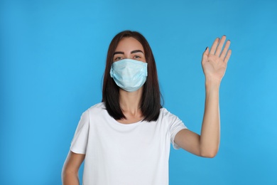 Photo of Young woman in protective mask showing hello gesture on light blue background. Keeping social distance during coronavirus pandemic