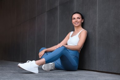 Photo of Smiling woman in sportswear sitting near dark grey wall outdoors