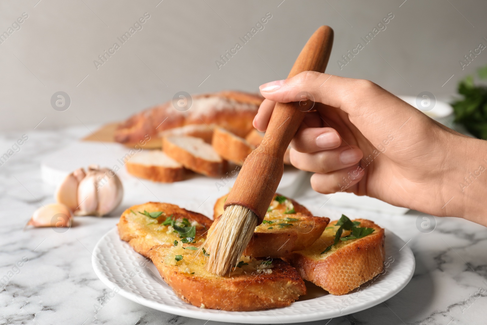 Photo of Woman brushing slices of delicious toasted bread with garlic and herbs on white marble table, closeup