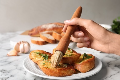 Photo of Woman brushing slices of delicious toasted bread with garlic and herbs on white marble table, closeup