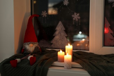 Gnome, burning candles and plaid near window with beautiful drawing at home. Christmas decor