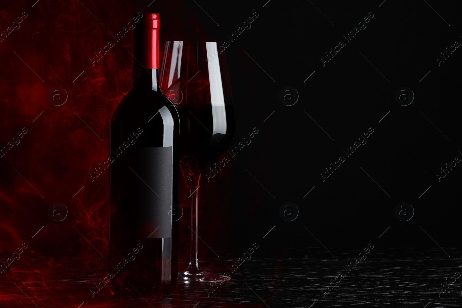 Photo of Tasty wine in glass and bottle in red lights on textured table against black background, space for text