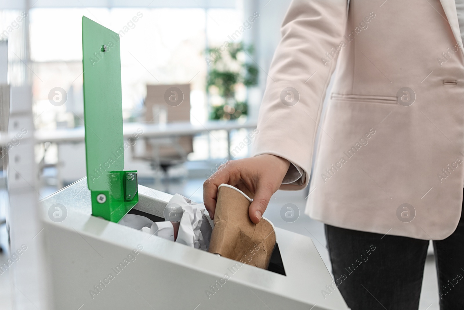 Photo of Woman putting used paper cup into trash bin in modern office, closeup. Waste recycling