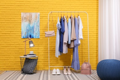 Rack with different stylish clothes and shoes near yellow brick wall in room