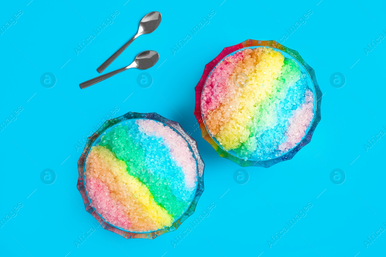 Photo of Rainbow shaving ice in glass dessert bowls and spoons on light blue background, flat lay