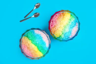 Photo of Rainbow shaving ice in glass dessert bowls and spoons on light blue background, flat lay