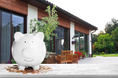 Image of Piggy bank and coins on stone surface and beautiful view of house, space for text. Mortgage concept