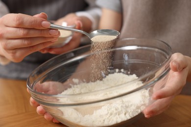 Photo of Making bread. Mother and her daughter putting flour and dry yeast into bowl at wooden table in kitchen, closeup