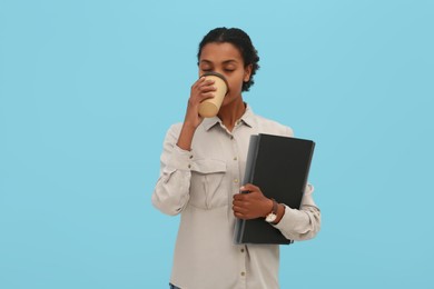 Photo of African American intern with folders drinking coffee on light blue background