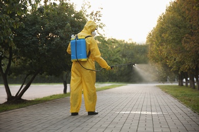 Photo of Person in hazmat suit disinfecting street pavement with sprayer, back view. Surface treatment during coronavirus pandemic