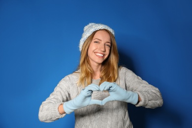 Image of Happy young woman in warm sweater making heart with hands on blue background