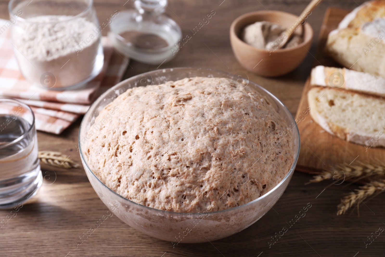 Photo of Fresh sourdough in bowl, water, spikes and bread on wooden table, closeup