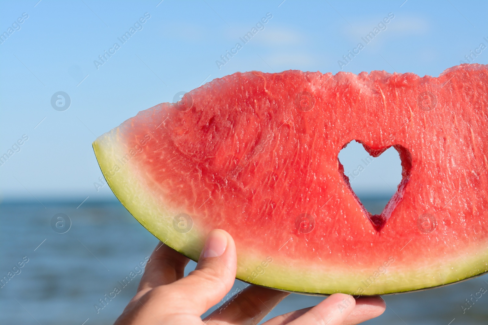 Photo of Child holding slice of watermelon with heart shaped hole near sea, closeup