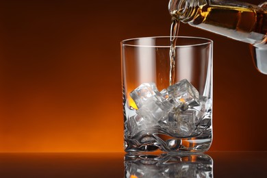 Pouring whiskey from bottle into glass with ice cubes at table against color background, space for text