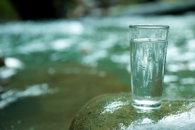 Photo of Glass of fresh water on stone near stream. Space for text