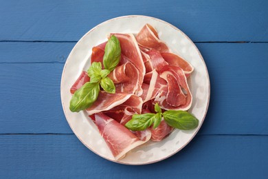 Photo of Slices of tasty cured ham and basil on blue wooden table, top view