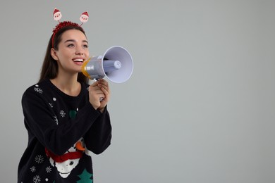Photo of Happy young woman in Christmas sweater and Santa headband shouting in megaphone on grey background. Space for text
