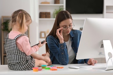 Photo of Woman working remotely at home. Tired mother using computer while her daughter playing with phone. Child sitting on desk