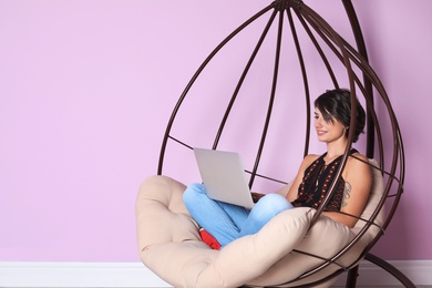 Young woman with modern laptop sitting in hanging chair near color wall