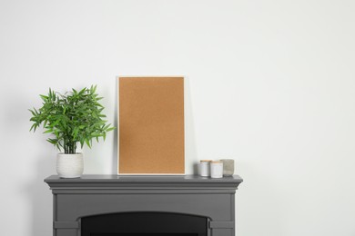 Empty frame, candles and potted houseplant on fireplace near white wall indoors, space for text. Interior design